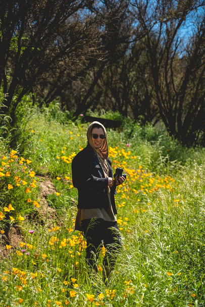 Aventure en montagne ensoleillée : un jeune photographe en tenue sombre explore une prairie ensoleillée parsemée de marguerites jaunes - Photo, image