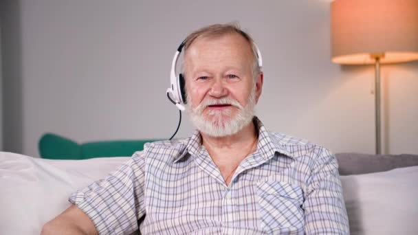 portrait d'un homme âgé parlant à l'aide d'un casque par vidéoconférence sur un ordinateur portable en famille assis sur le canapé dans une pièce confortable, regardant la caméra - Séquence, vidéo