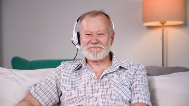 videollamada en línea, hombre viejo alegre moderno hablando por webcam con auriculares mientras se relaja en la habitación en el sofá, mirando a la cámara - Imágenes, Vídeo