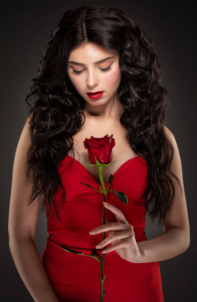 Μια λεπτή μελαχρινή γυναίκα με μακριά σγουρά μαλλιά και όμορφο μακιγιάζ, φοράει ένα κόκκινο φόρεμα, κρατά ένα τριαντάφυλλο. - Φωτογραφία, εικόνα