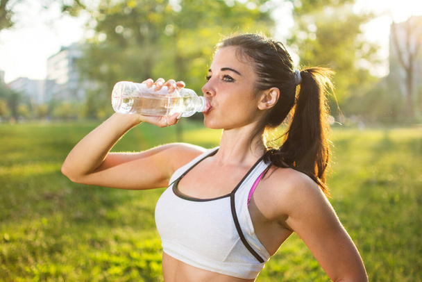 Joven mujer sonriente bebiendo agua de la botella después de ejercicio deportivo al aire libre al atardecer - Foto, imagen