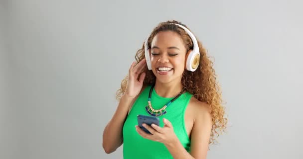 Happy woman, phone and dancing with headphones to music or podcast on a gray studio background. Excited female person or model listening and enjoying audio sound on mobile smartphone or mockup space. - Footage, Video