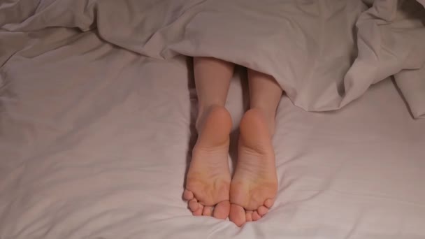 女性の足は白いシートの上の毛布の下で夜に眠っている. 女性の女の子は裸足のベッドルームでベッドで寝る - 映像、動画