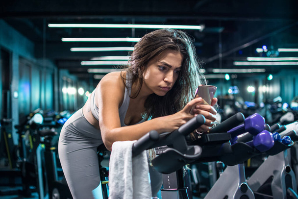 Attraktive junge Frau telefoniert während Trainingspause auf dem Indoor-Fahrrad - Foto, Bild