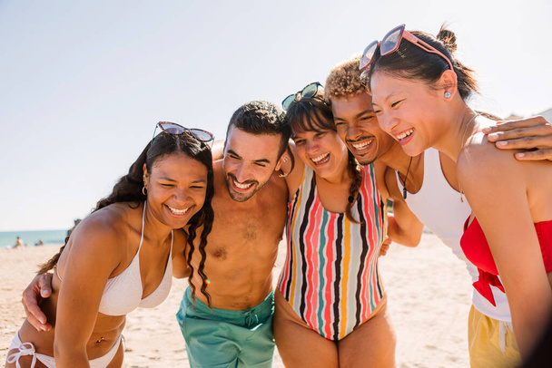 Молодая группа людей весело обнимается на пляже, наслаждаясь летним отпуском вместе. Концепция дружбы и отдыха. - Фото, изображение