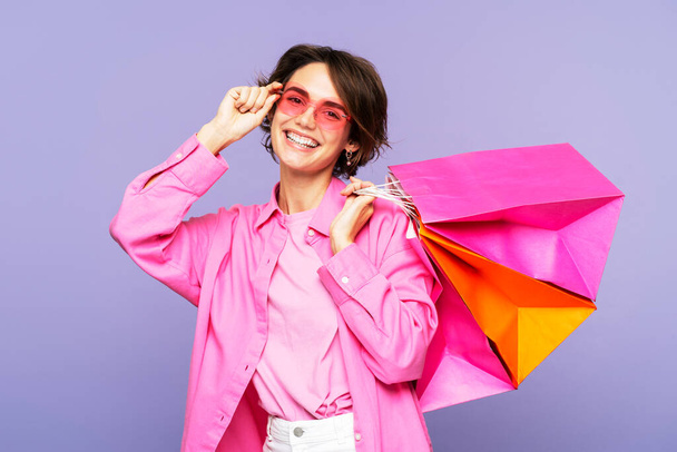 Krásná, usmívající se žena ve slunečních brýlích a růžových šatech, držící několik papírových nákupních tašek, stojící izolovaně na šeříkovém pozadí a hledící přímo do kamery. Nákupní koncept - Fotografie, Obrázek