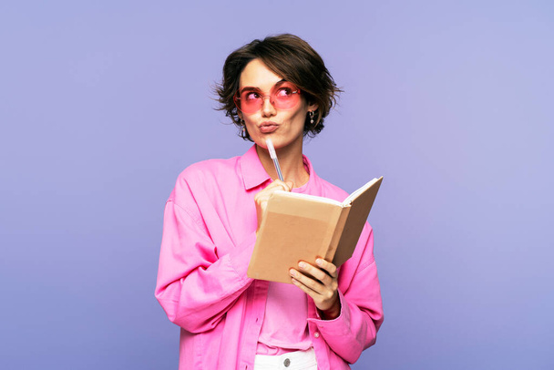 Picture of an adorable young stylish woman, wearing sunglasses, with a serious facial expression, holding a pen and diary, standing isolated on a lilac background, looking away - Photo, Image