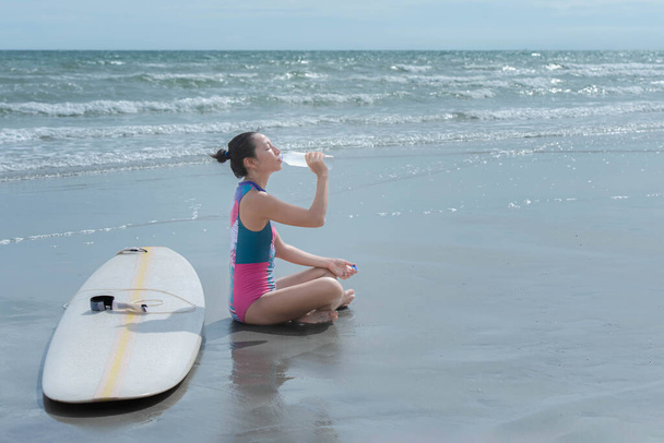 Mladá Asiatka sedící na pláži Pila vodu z láhve, aby uhasila žízeň. po cvičení Koncept hraní surfařských sportů pro zdraví - Fotografie, Obrázek