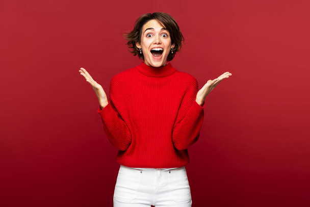Aantrekkelijke jonge vrolijke vrouw in een portret, verrast en gebarend met wijd open mond dramatisch, geïsoleerd op een rode achtergrond, kijkend naar de camera - Foto, afbeelding