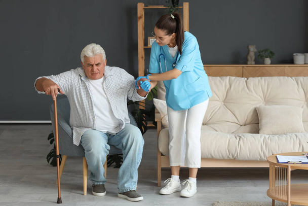 Verpleegster helpen senior man met stok om op te staan uit fauteuil thuis - Foto, afbeelding