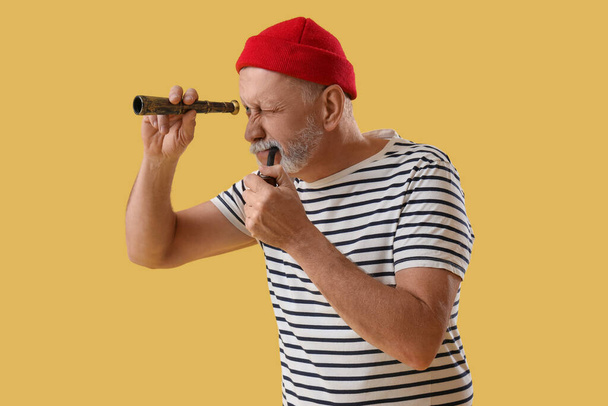 Mature sailor with smoking pipe looking through spyglass on yellow background - Photo, Image