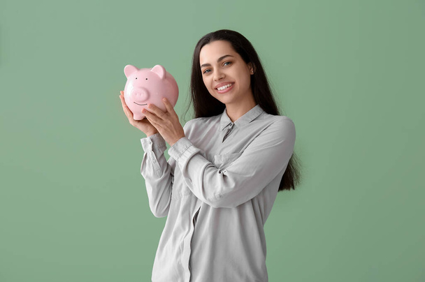 Beautiful young happy woman with piggy bank on green background - Photo, Image