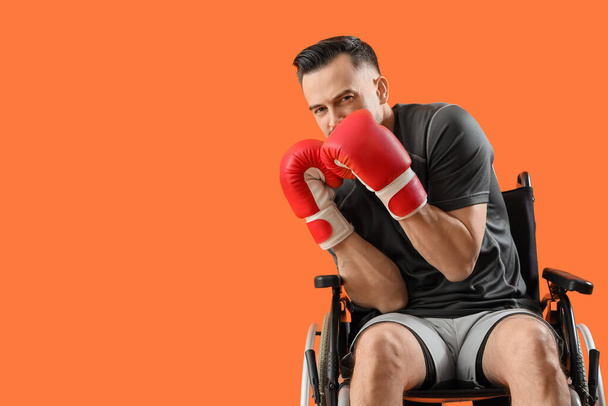 Спортивний молодий чоловік в інвалідному кріслі з боксерськими рукавичками на помаранчевому фоні - Фото, зображення