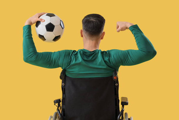 Mannelijke voetballer in rolstoel met bal met spieren op gele achtergrond, achteraanzicht - Foto, afbeelding
