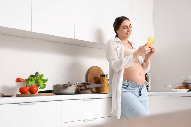 Νεαρή έγκυος γυναίκα που χρησιμοποιεί κινητό τηλέφωνο ενώ μαγειρεύει στην κουζίνα - Φωτογραφία, εικόνα