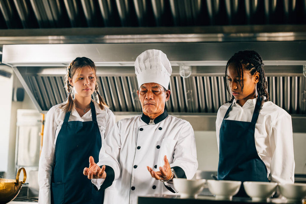 Profesyonel Asyalı kıdemli şef, restoran mutfağındaki bir atölyede takım çalışması öğrenme ve not alma üzerine çalışan çeşitli öğrencilere rehberlik ediyor. Eğitim ortamı. Gıda Eğitimi - Fotoğraf, Görsel