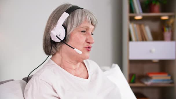 σύγχρονοι συνταξιούχοι, χαρούμενη ηλικιωμένη γυναίκα με ακουστικά που μιλάει σε μια βιντεοκλήση, ενώ κάθεται στον καναπέ στο δωμάτιο - Πλάνα, βίντεο