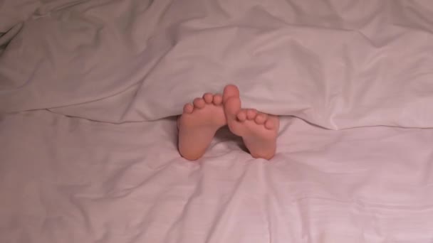 Çocuk bacakları, geceleri battaniyenin altında parmaklarıyla dans eder ve eğlenir. Çıplak ayaklı mutlu kız bebek geceleri yatak odasındaki rahat yatakta uyumuyor. - Video, Çekim