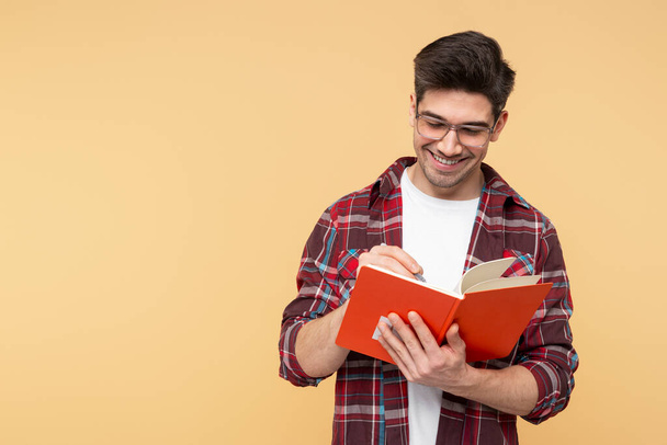 Portret uśmiechniętego, mądrego hipstera w okularach, trzymającego w sobie książkę i piszącego w niej, pozującego na żółtym tle, kopiującego miejsce na reklamę - Zdjęcie, obraz