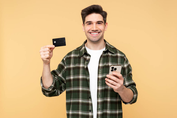 Фото веселого человека с широкой улыбкой, держащего мобильный телефон и демонстрирующего кредитную карту, изолированную на желтом фоне и смотрящего прямо в камеру - Фото, изображение