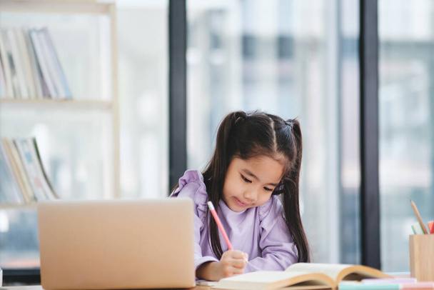 Een jong meisje zit achter een bureau met een laptop en een boek. Ze schrijft op de laptop en richt zich op haar werk. - Foto, afbeelding