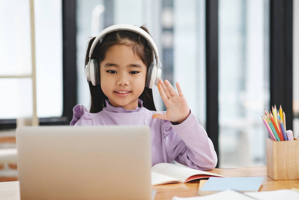 Een jong meisje met een koptelefoon zwaait naar de camera terwijl ze achter een bureau zit met een laptop en een boek - Foto, afbeelding
