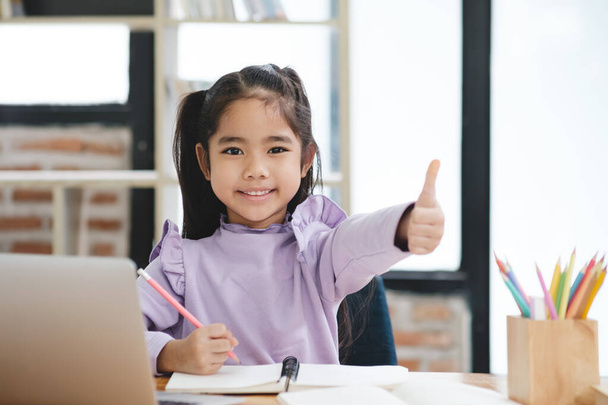 Een jong meisje zit achter een bureau met een laptop en een notitieboekje. Ze glimlacht en geeft een duim op. De scène suggereert dat ze gelukkig is en geniet van haar werk of activiteit - Foto, afbeelding