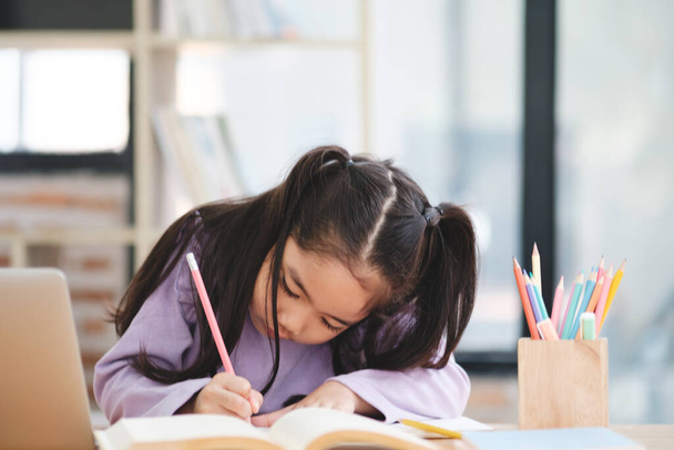 Młoda dziewczyna siedzi przy biurku z ołówkiem i książką. Pisze w książce. - Zdjęcie, obraz