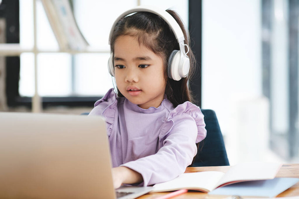Une jeune fille est assise à un bureau avec un ordinateur portable et un cahier. Elle porte des écouteurs et se concentre sur son travail. La scène suggère qu'elle est engagée dans une tâche qui nécessite de la concentration - Photo, image