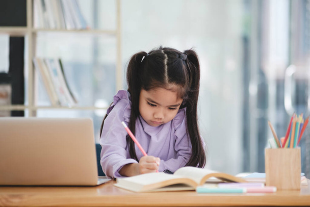 Une jeune fille est assise à un bureau avec un ordinateur portable et un livre. Elle écrit avec un crayon et se concentre sur son travail. La scène suggère une atmosphère calme et studieuse - Photo, image