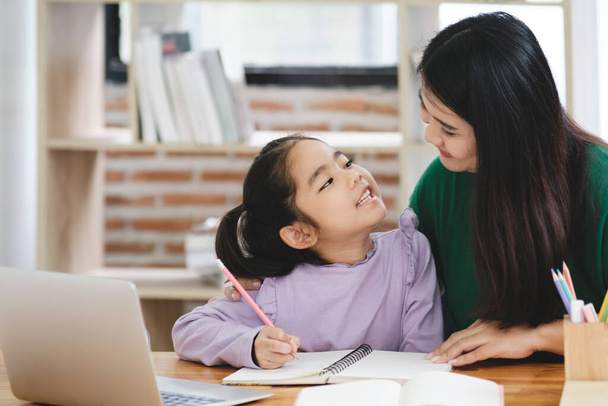 Žena pomáhá mladé dívce s domácím úkolem. Dívka má na sobě fialovou košili a sedí u stolu s notebookem a zápisníkem. Ta žena drží děvčata za ruku. - Fotografie, Obrázek