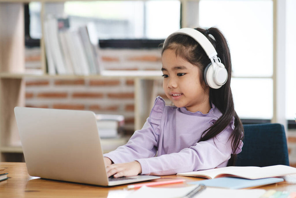 Genç bir kız dizüstü bilgisayarı ve kulaklığı olan bir masada oturuyor. Gülümsüyor ve işinden zevk alıyor. - Fotoğraf, Görsel