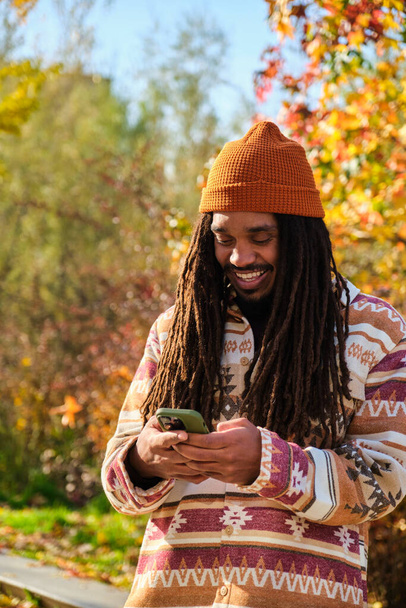 jovem afro-americano com dreadlocks feliz usando o smartphone em um dia ensolarado de outono. - Foto, Imagem