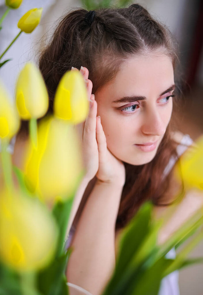 Una joven con el pelo largo se sienta en una mesa de madera blanca con un jarrón de tulipanes amarillos. - Foto, Imagen
