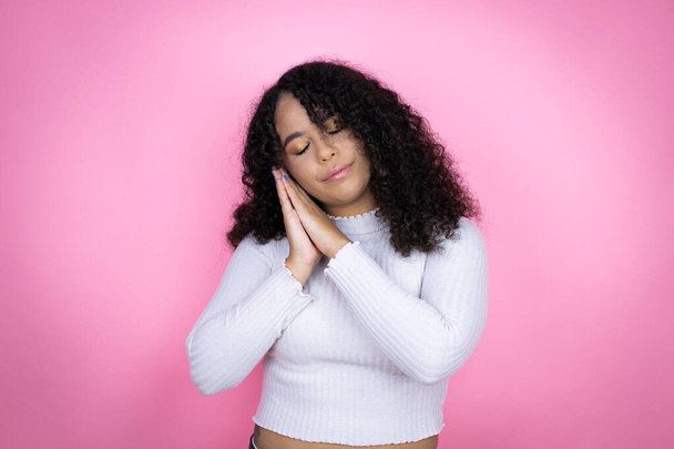 Αφροαμερικανή γυναίκα φορώντας casual πουλόβερ πάνω από ροζ φόντο κοιμάται κουρασμένος όνειρα και ποζάρουν με τα χέρια μαζί, ενώ χαμογελά με κλειστά μάτια. - Φωτογραφία, εικόνα
