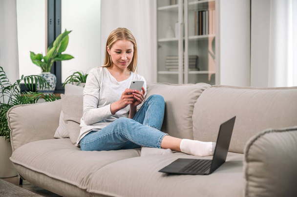 Šťastná žena odpočívat na pohodlné pohovce a pomocí moderních gadgets s internetem doma. Volně stojící psaní zprávy na smartphone pro pracovní chat, sedí v blízkosti notebooku a sledovat online video - Fotografie, Obrázek