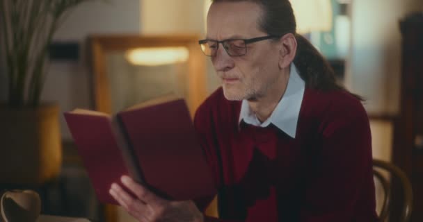 Gentiluomo anziano immerso nella lettura notturna, affascinato da un libro sotto una luce soffusa e soffusa nel comfort di casa - Filmati, video