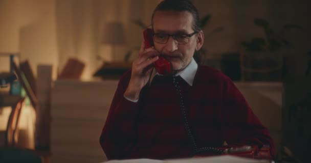 Anciano caballero se involucra en una conversación nostálgica en un teléfono vintage bajo el ambiente tranquilo de la noche - Imágenes, Vídeo