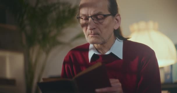 Ηλικιωμένος κύριος βυθισμένος στη νυχτερινή ανάγνωση, γοητευμένος από ένα βιβλίο κάτω από μαλακό, αμυδρό φως στην άνεση του σπιτιού - Πλάνα, βίντεο