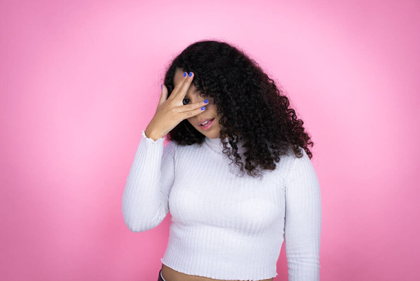 ピンクの背景にカジュアルなセーターを身に着けているアフリカ系アメリカ人女性は,恥ずかしい表情で指を通して見て,顔と目をつぶります - 写真・画像