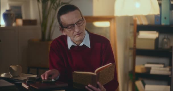Gentiluomo anziano immerso nella lettura notturna, affascinato da un libro sotto una luce soffusa e soffusa nel comfort di casa - Filmati, video