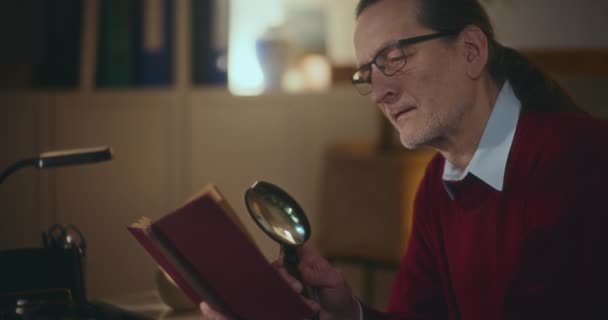 Signore anziano assorto nella lettura notturna, utilizzando una lente d'ingrandimento per approfondire il suo libro, alla ricerca di conoscenza sotto la luce fioca - Filmati, video