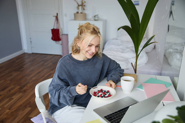 Obraz uśmiechniętej młodej kobiety oglądającej filmy na laptopie podczas jedzenia śniadania i picia herbaty, patrzącej na ekran, siedzącej w swoim pokoju. - Zdjęcie, obraz