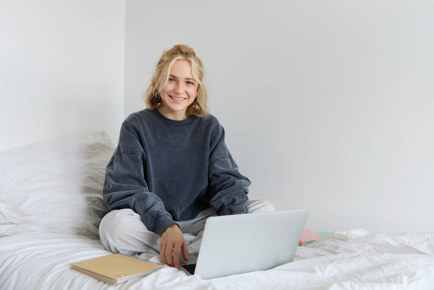 Ritratto di giovane donna sorridente che studia nel suo letto, lavora da casa in camera da letto, seduta con laptop e quaderni in posa di loto, sembra felice e rilassata. - Foto, immagini