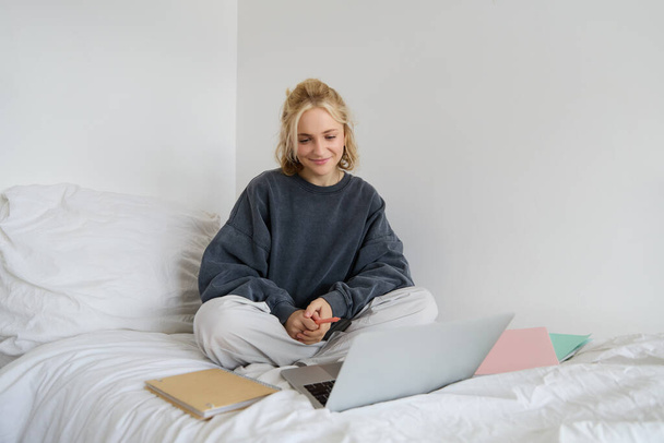 Портрет молодой студентки, женщины, обучающейся онлайн, обучающейся на ноутбуке, сидящей на кровати с ноутбуком и смотрящей на экран, видеочаты, подключающейся к саморазвивающемуся курсу. - Фото, изображение