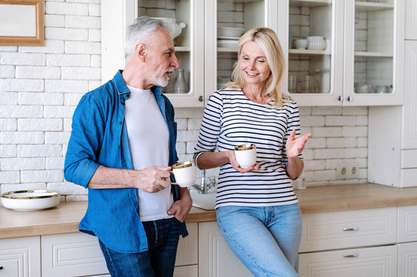 Volwassen echtpaar genieten van 's morgens koffie in de moderne keuken van nieuw appartement. Vrouw en man staan en praten in een modern huis. Samen tijd doorbrengen. - Foto, afbeelding