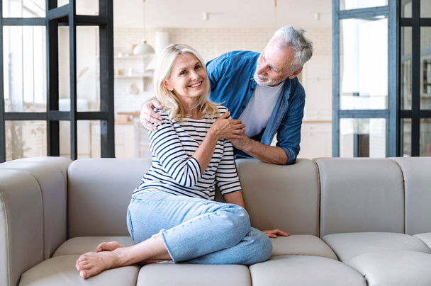 Зрелый мужчина обнимается и смотрит на счастливую улыбающуюся жену, сидящую на диване. Наслаждайтесь свободным временем в светлом интерьере гостиной. Семейная любовь, отношения, поддержка и уход дома - Фото, изображение