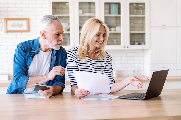 Paying invoices online. Smiling mature woman check utility bills for water supply, gas and electricity on laptop and showing infomation for man. Couple filling tax forms together. - Foto, Bild
