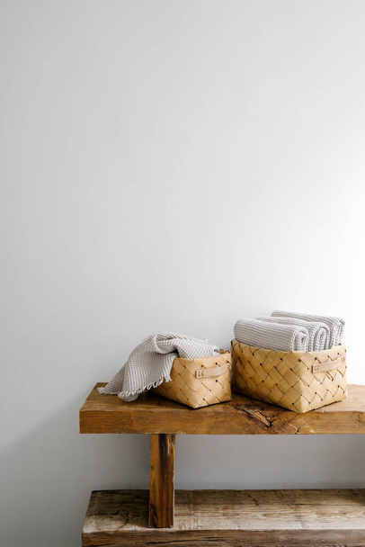 Vertikale Aufnahme von geflochtenen Körben mit sauberen Handtüchern aus frischer Baumwolle auf einer Holzbank vor einer weißen Wand im Badezimmer der Wohnung. Minimalistisches Interieur mit Wohnkultur. Kopierraum - Foto, Bild