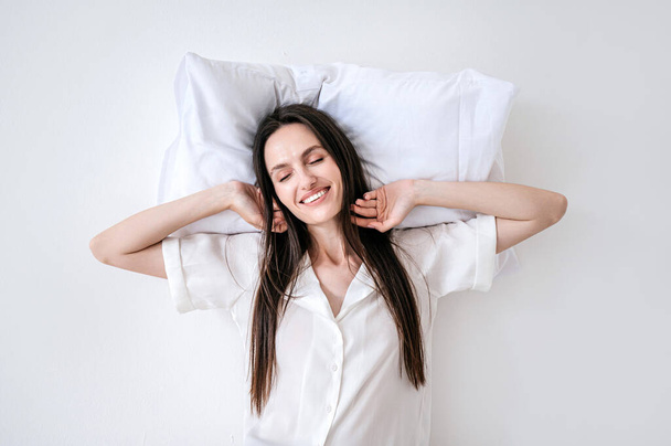 Pijamalı mutlu bir kadın, rahat bir yastıkta uyuyor, uyanıyor, ellerini kaldırıyor, esniyor ve gülümsüyor beyaz arka planda kapalı gözlerle. - Fotoğraf, Görsel